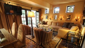 Un salon chaleureux avec un ensemble de canapés, une table basse et un tapis à motifs. On y trouve plusieurs fenêtres avec des rideaux, un téléviseur à écran plat et des lampes fixées au mur.