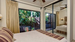 Une chambre avec un lit, une grande porte-fenêtre coulissante donnant sur un jardin, un ventilateur de plafond et une table de chevet avec une lampe.