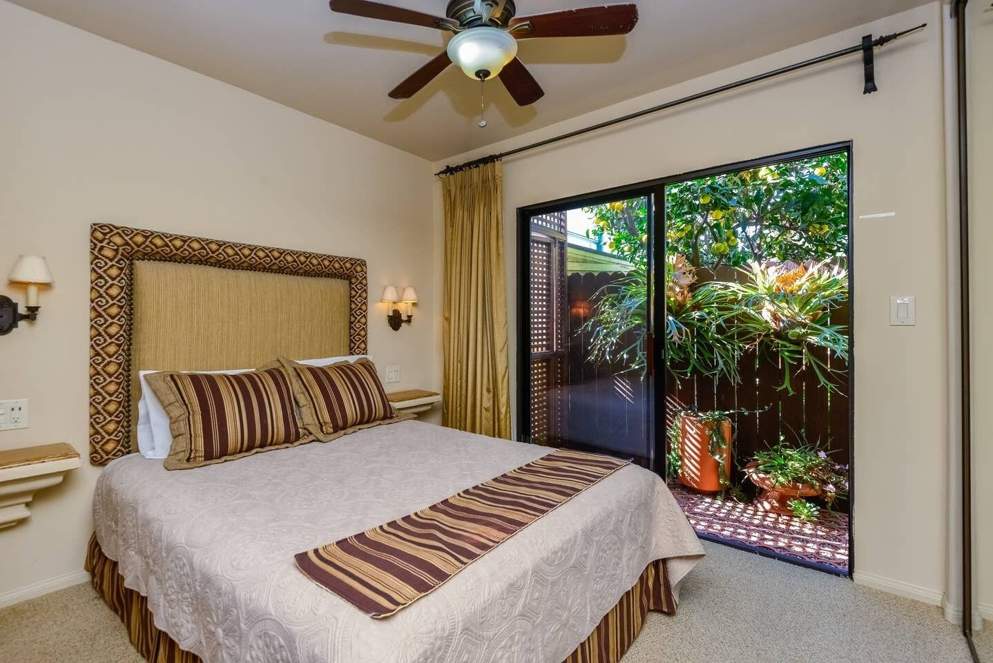 A bedroom with a bed, a ceiling fan, a nightstand, a wall-mounted lamp, and a view of a garden through a sliding door.