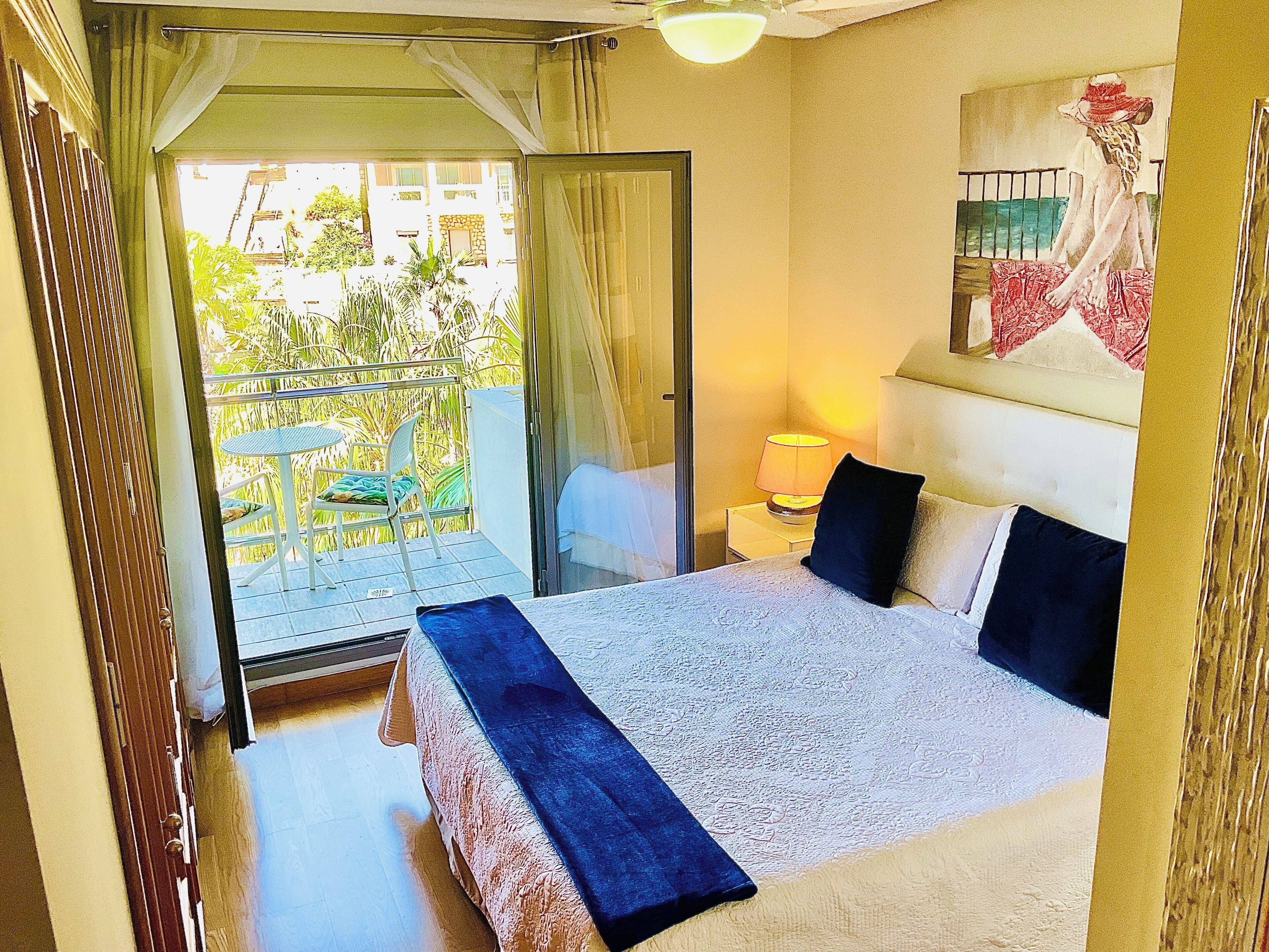 Ein Hotelzimmer mit Bett, Nachttisch, Lampe, einem Fenster mit Vorhängen und Blick auf den Außenbalkon.