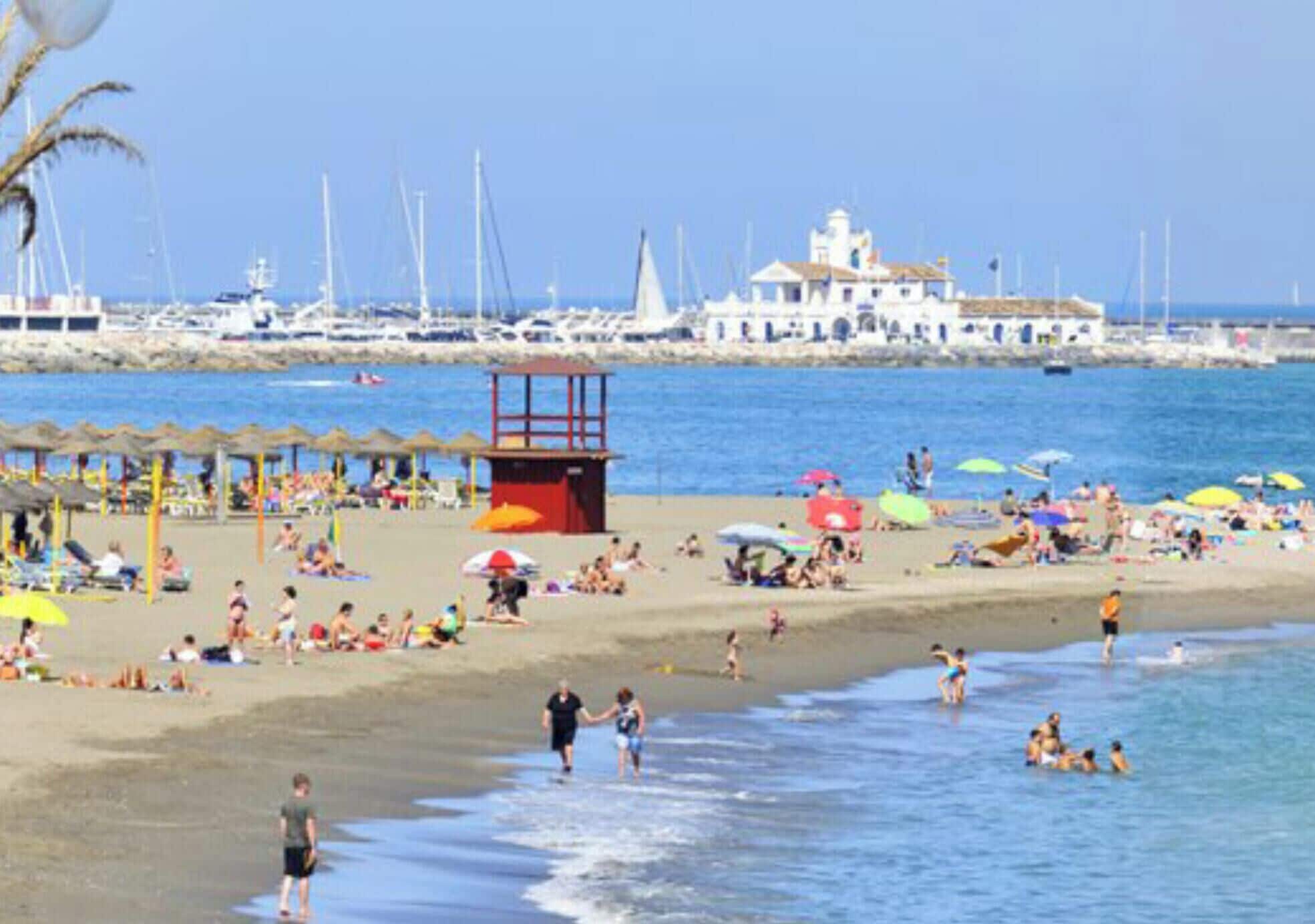 Une plage où des gens se prélassent au soleil et se baignent, une tour de surveillance des sauveteurs et des bateaux en arrière-plan.