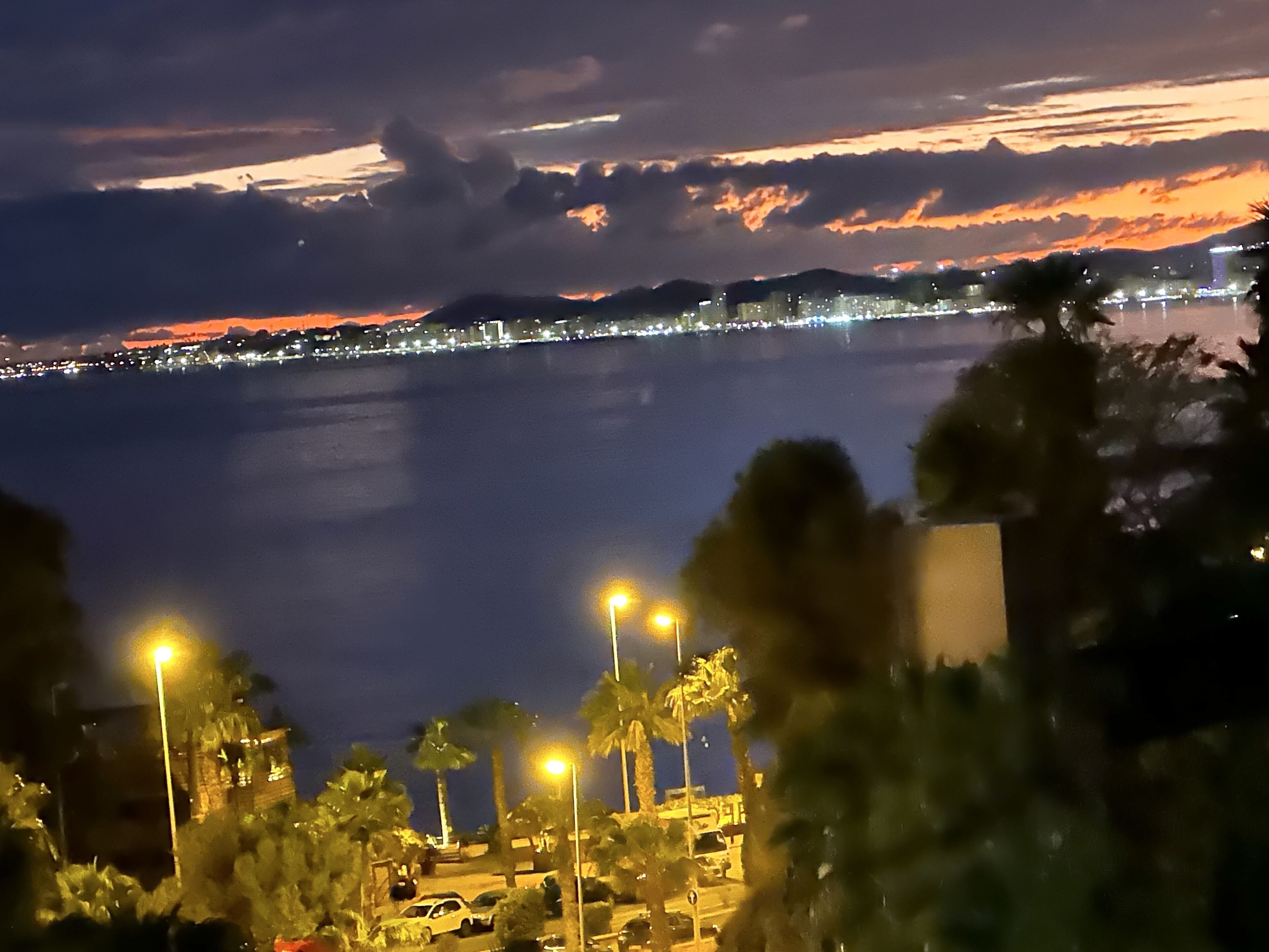 A nighttime view of a coastal city with a body of water, street lights, and distant city lights.