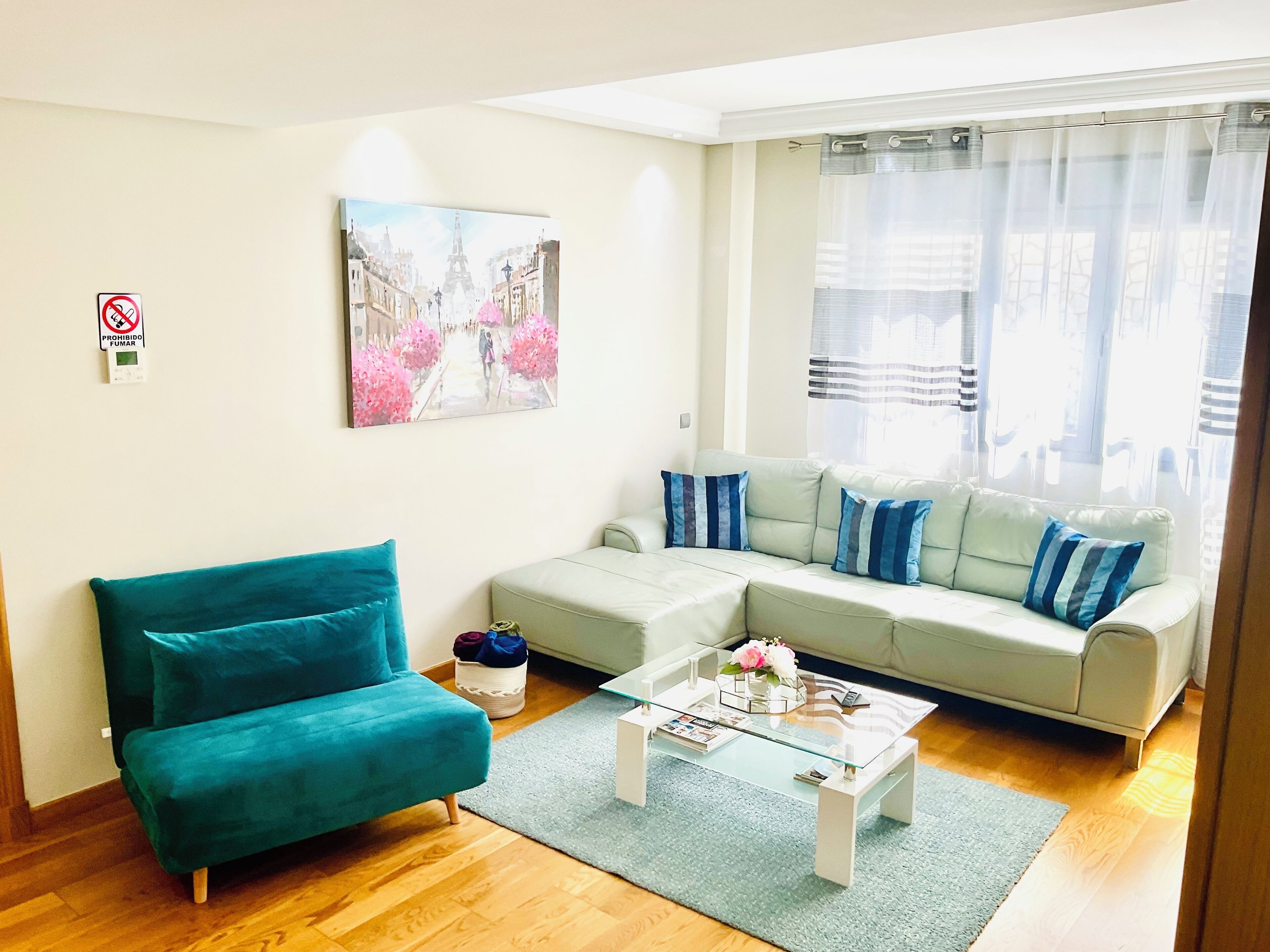 A living room with a teal sofa, a light-colored sectional sofa, a glass coffee table, and a painting of a city street.