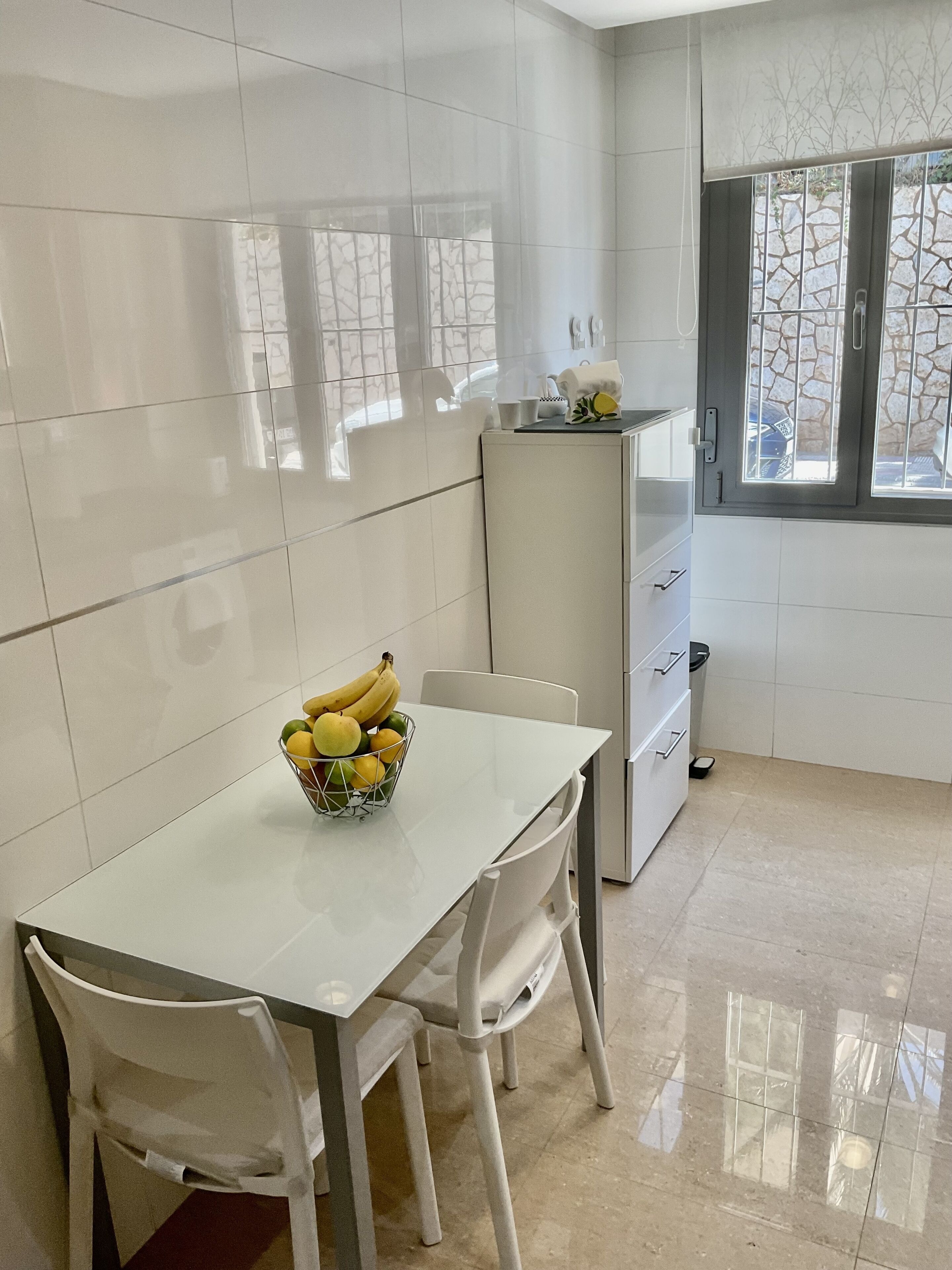 Een moderne keuken met witte tegels, een eettafel met stoelen, een fruitschaal en een raam met uitzicht op een stenen muur.
