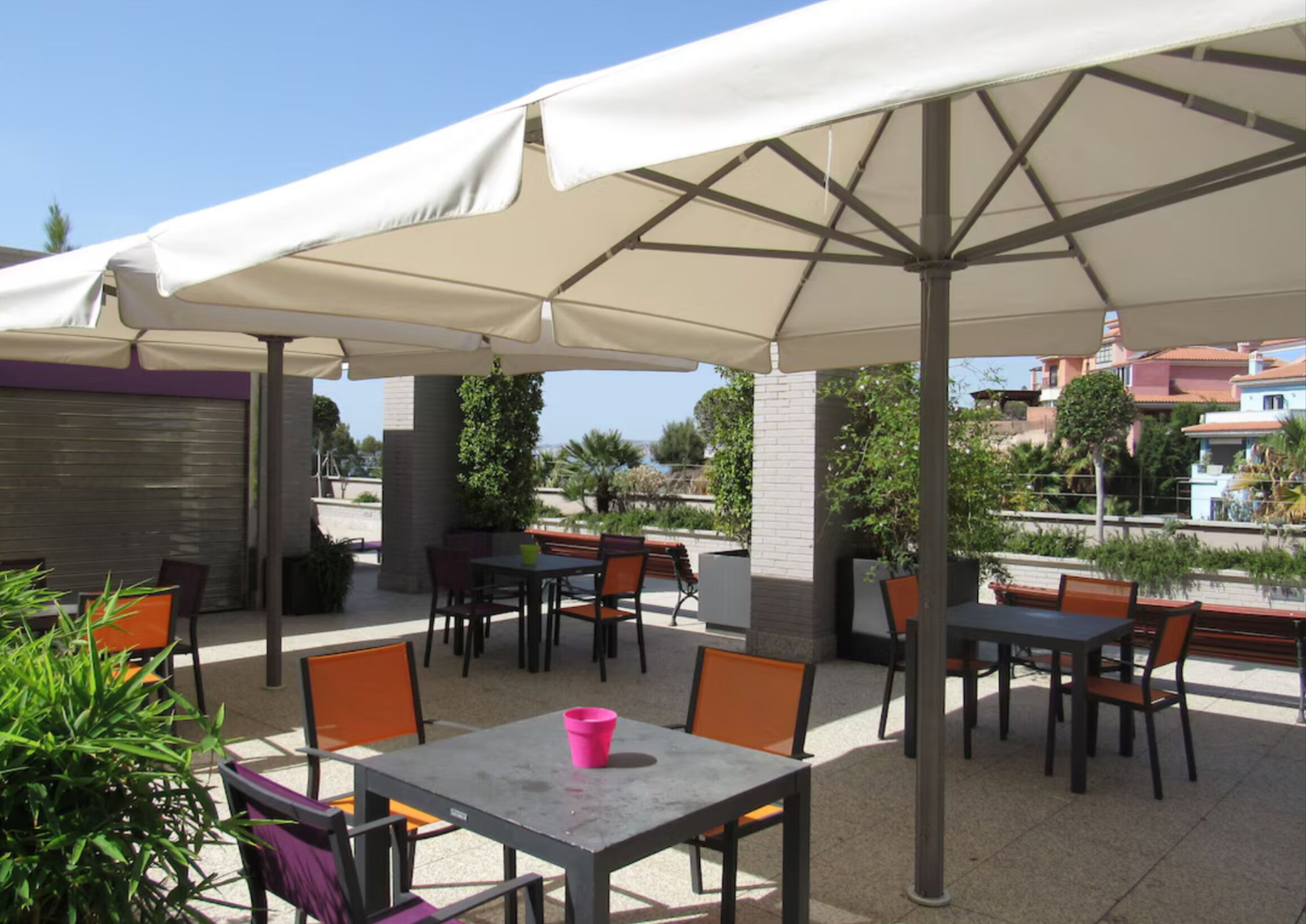 Een terras buiten met tafels en stoelen onder grote parasols, omgeven door groen en gebouwen op de achtergrond.