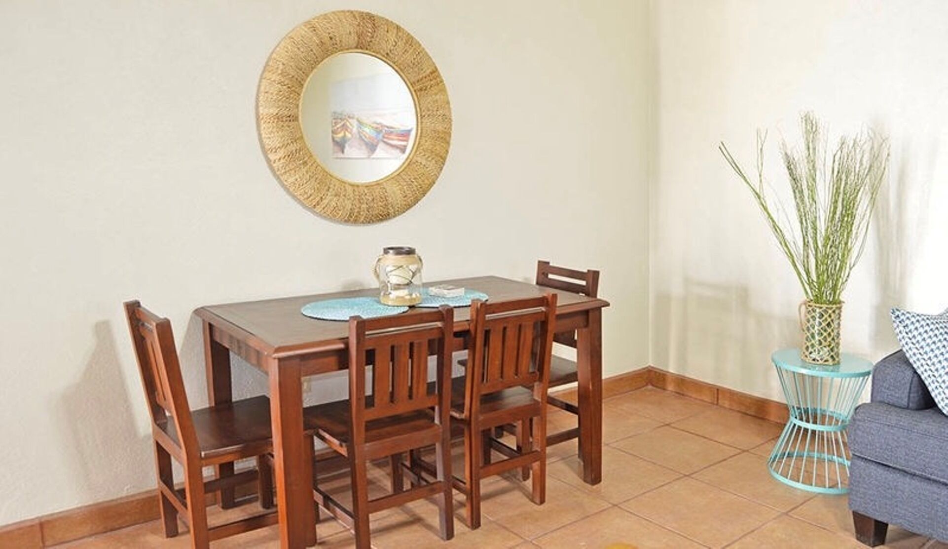 A dining area with a wooden table and chairs, a round mirror on the wall, and a small side table with a vase of plants.