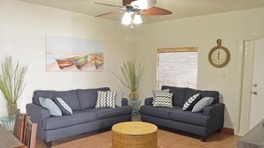 Ein Wohnzimmer mit zwei grauen Sofas, einem Deckenventilator, einem Gemälde von Booten, einem Fenster mit Jalousien und einer Tür mit einer Uhr.