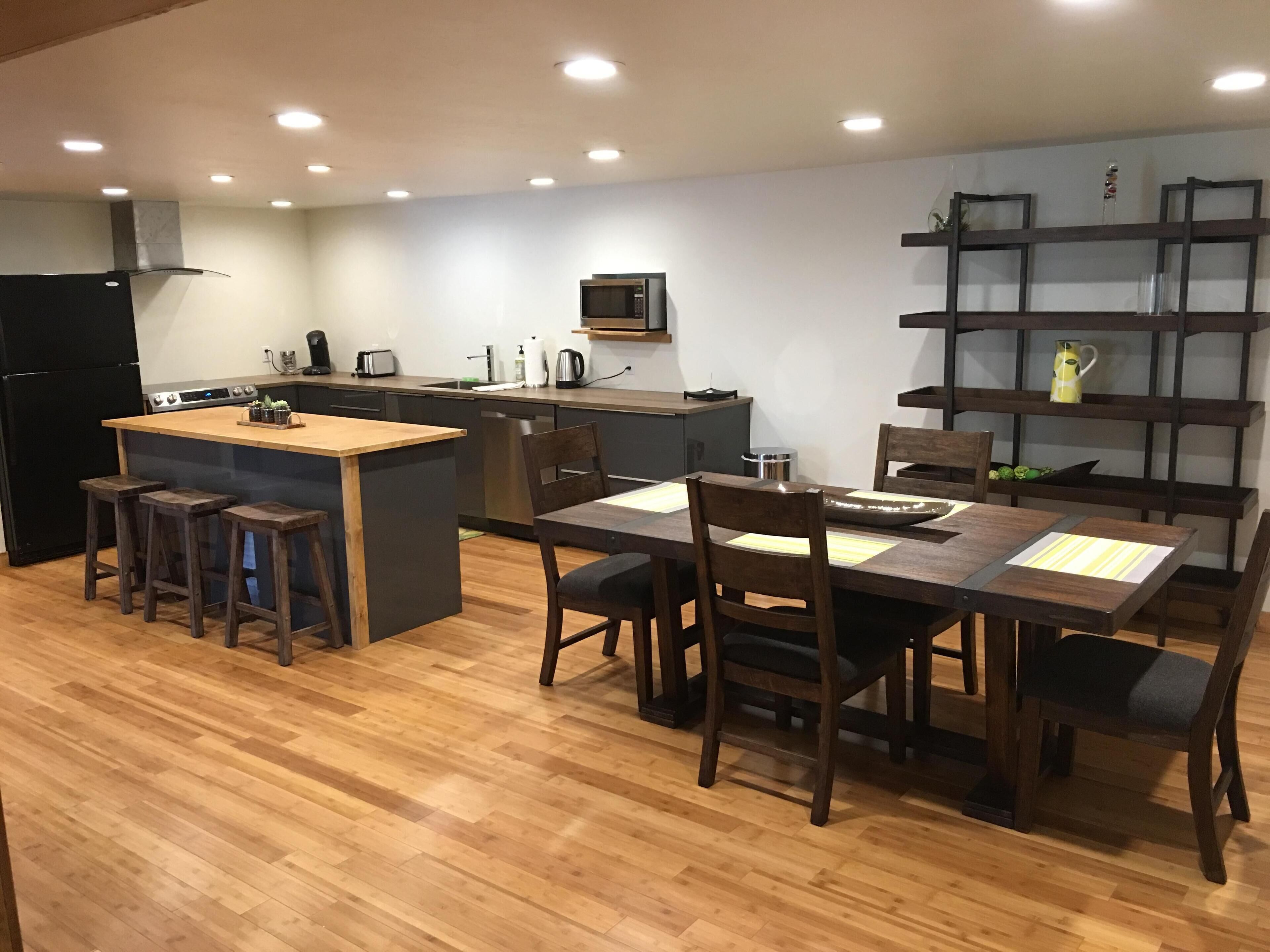 A modern kitchen with a central island, wooden stools, a dining table with chairs, and built-in shelving.