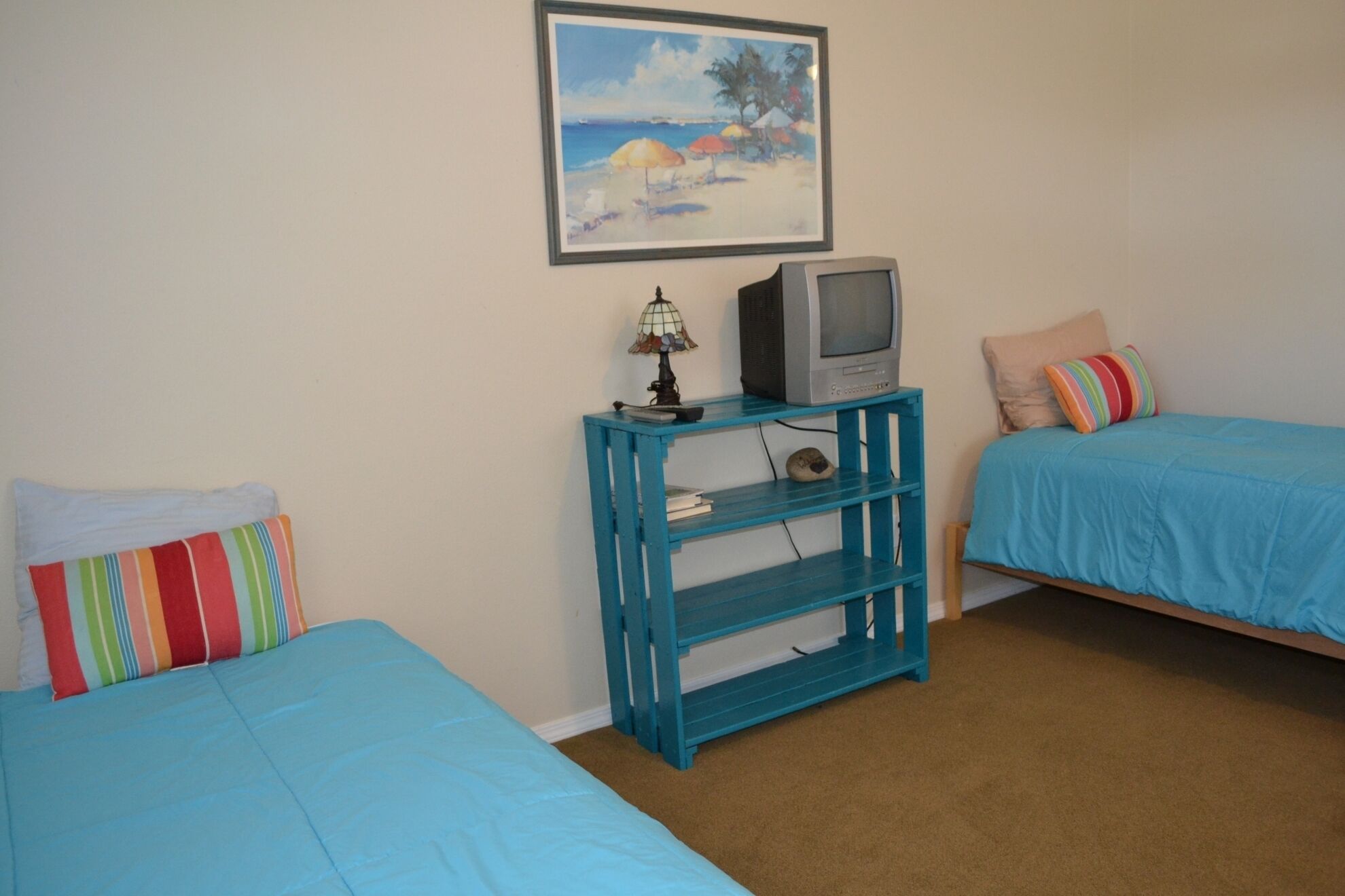 A bedroom with two beds, a TV on a blue shelf, and a beach painting on the wall.