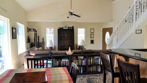 Ein geräumiges Wohnzimmer mit hohen Decken, Kamin, Bücherregal, Essbereich mit Tisch und Stühlen sowie einer Treppe mit Geländer.
