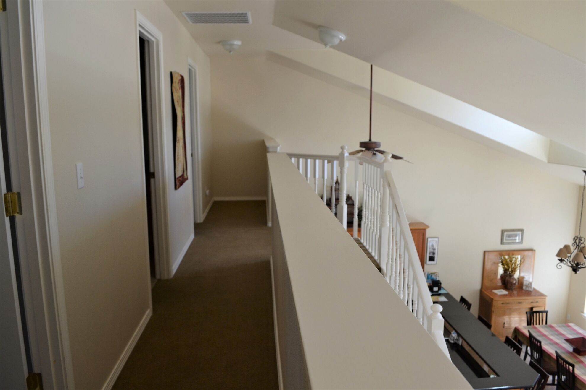 A hallway with a carpeted floor, white walls, and a white railing leading to a dining area with a table and chairs.