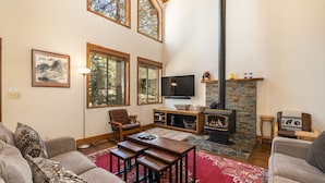 A cozy living room with a fireplace, a television, and a large window overlooking a forest. - Creekside Cabin - Beautiful Dog Friendly Cabin in TD, Hiking and Biking Access (Truckee)