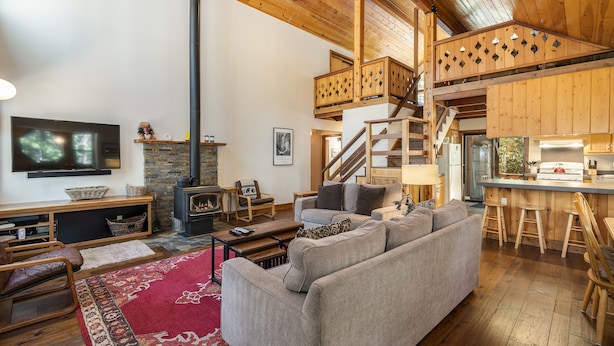 A spacious living area with a large sofa, a fireplace, a TV, and a kitchen area with stools. - Creekside Cabin - Beautiful Dog Friendly Cabin in TD, Hiking and Biking Access (Truckee)