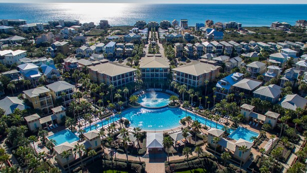 Een luchtbeeld van een resort met een centraal gelegen zwembad, omringd door villa's, palmbomen en uitzicht op de oceaan.
