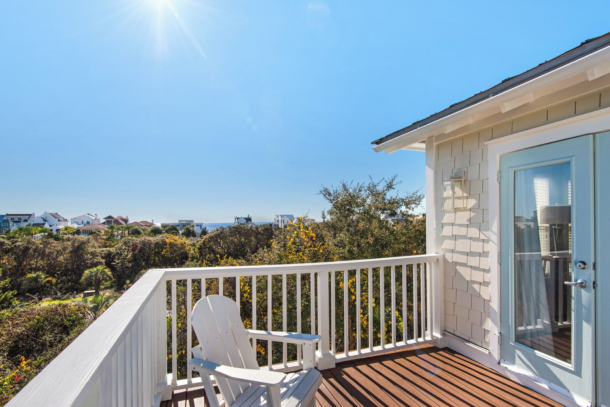 A balcony with a white chair, a wooden deck, and a view of the sea and other buildings.