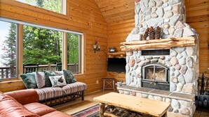 A cozy cabin interior with a stone fireplace, wooden walls, a sofa, and a large window overlooking greenery.