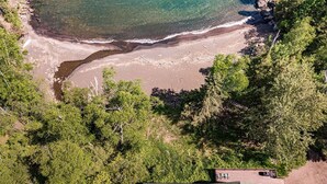An aerial view of a beach with clear water, surrounded by dense greenery and a house at the bottom.