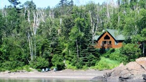 A cabin with a green roof, surrounded by dense forest, near a calm lake.