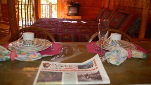 A wooden cabin interior with a bed, a television, and a dining table set with plates, glasses, and a newspaper. - Cozy Rustic Hot Tub Cabin w/Hike in River Access - SWB (Sevierville)