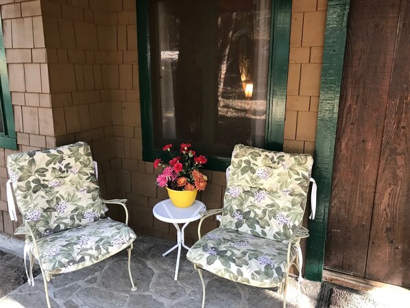 Two outdoor patio chairs with floral upholstery, a small round table with a yellow pot of flowers, and a brick wall with a window.