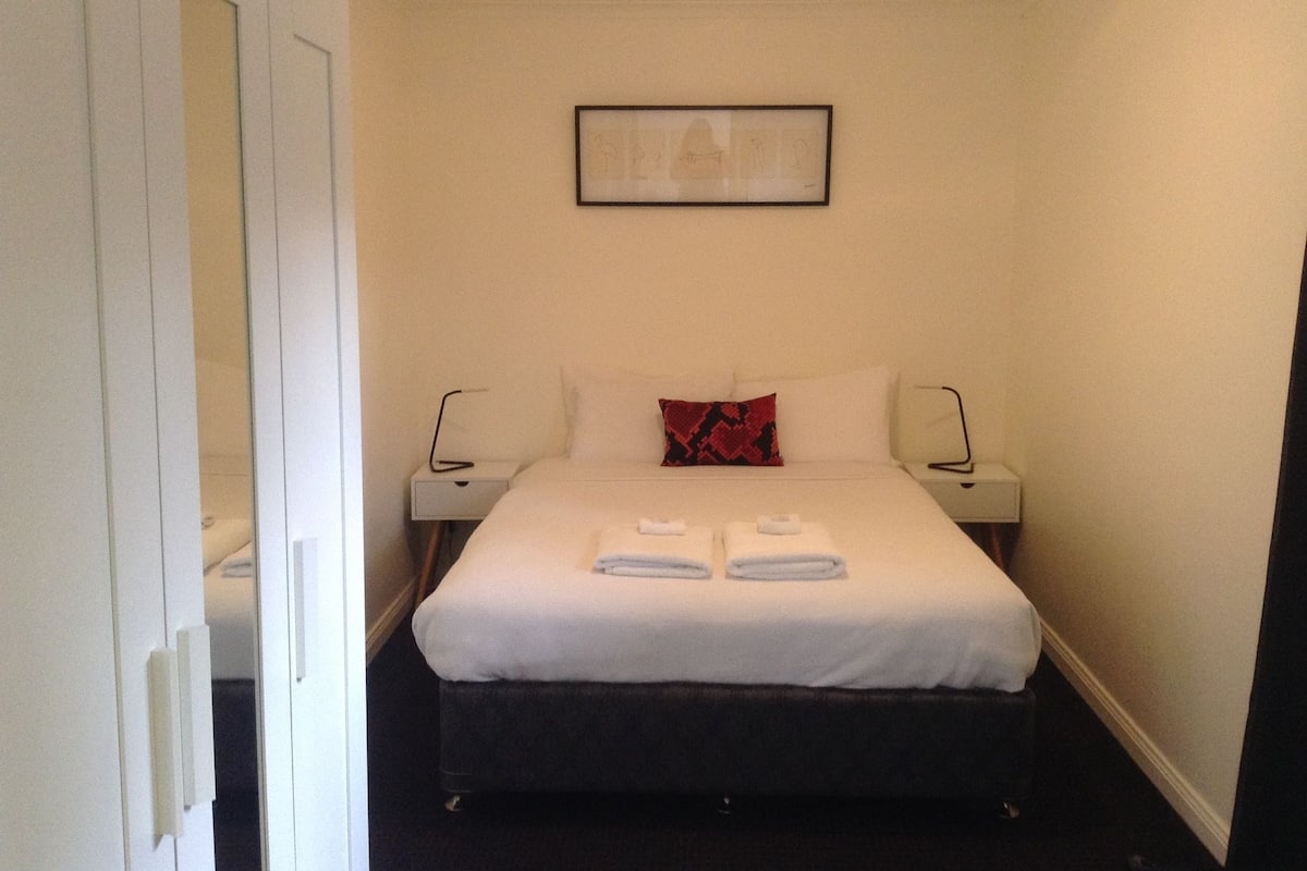 Habitación de hotel con cama, dos mesitas de noche, un buró, un cuadro en la pared y una puerta.