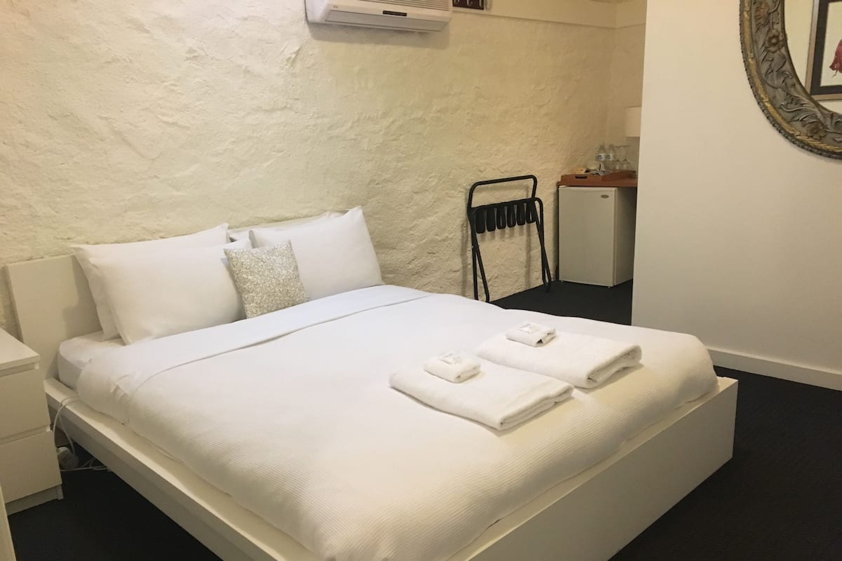 Una cama bien hecha con sábanas y toallas blancas, una mesita de noche, un aire acondicionado de pared y un pequeño aparador.