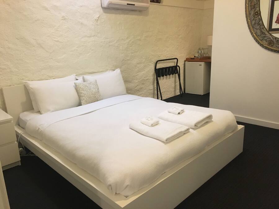 A neatly made bed with white linens and towels, a bedside table, a wall-mounted air conditioner, and a small bedside cabinet.