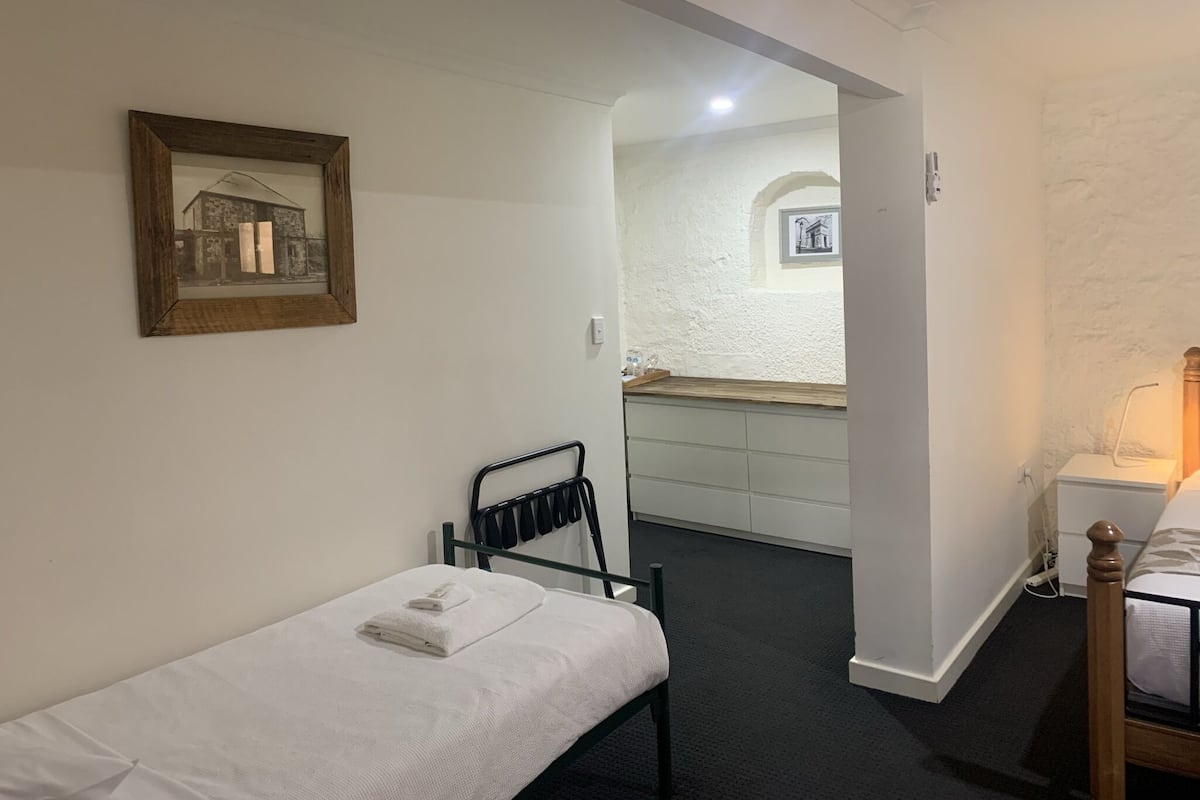 Habitación pequeña y limpia con una cama individual, una silla, un escritorio y un espejo. Hay un cuadro en la pared y una ventana pequeña que da a un edificio.