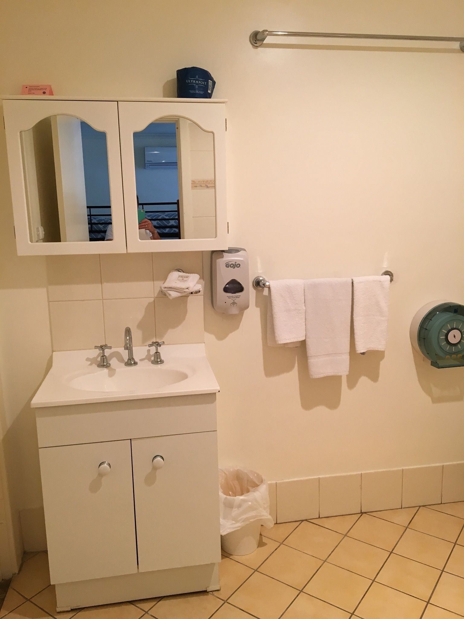 A bathroom with a white sink, two mirrors, a towel rack with two towels, a soap dispenser, and a trash bin.