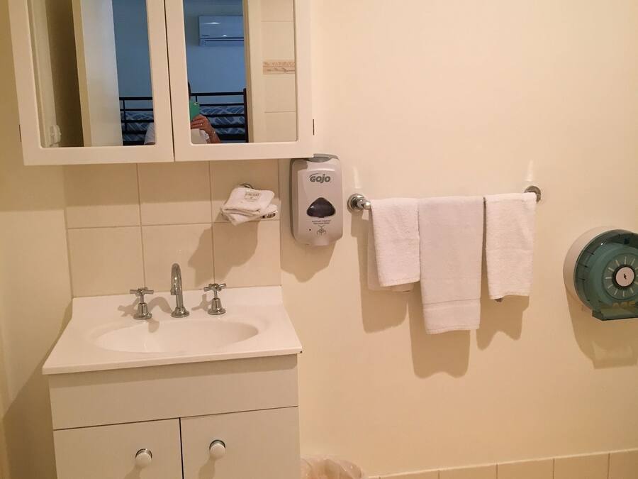 Une salle de bain avec un lavabo blanc, deux miroirs, un porte-serviettes avec deux serviettes, un distributeur de savon et une poubelle.