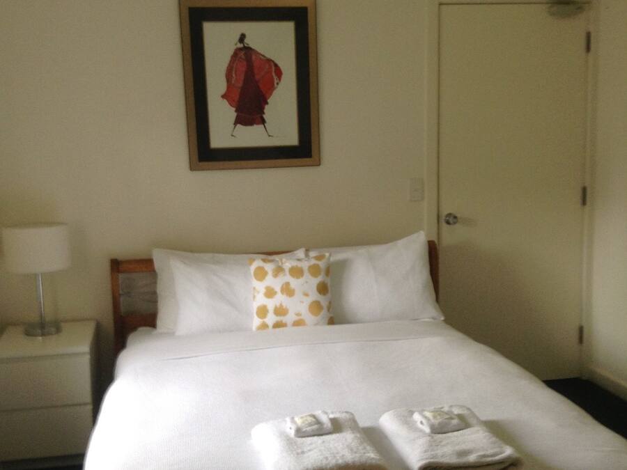 A neatly made bed with white linens and two folded towels, a nightstand with a lamp, a framed picture on the wall, and a closed door.