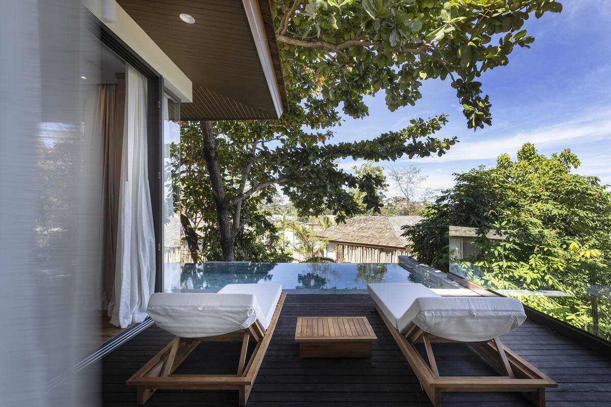A wooden deck with two sun loungers and a small table, overlooking a clear swimming pool and lush greenery.
