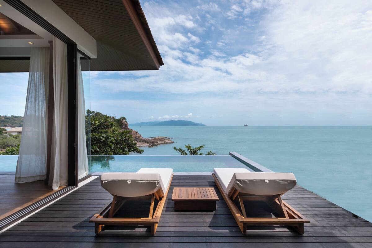 A wooden deck with two lounge chairs and a table, overlooking a clear blue sea and distant mountains.