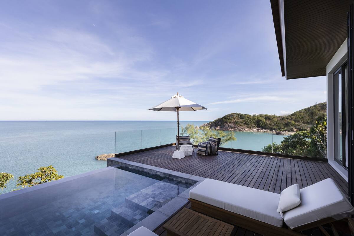 A modern outdoor deck with a glass infinity pool, wooden flooring, and a white lounge chair.
