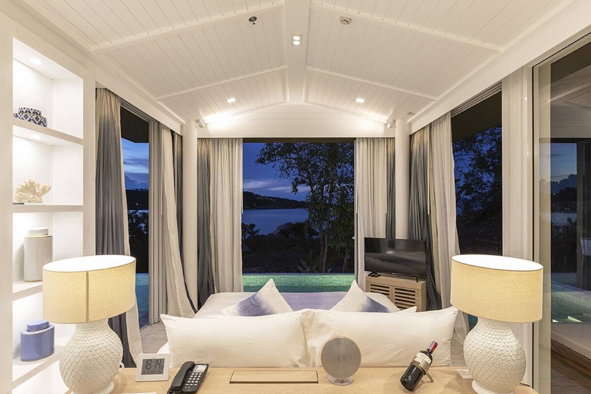 A bedroom with a bed, two bedside lamps, a TV, and a view of the outdoors.