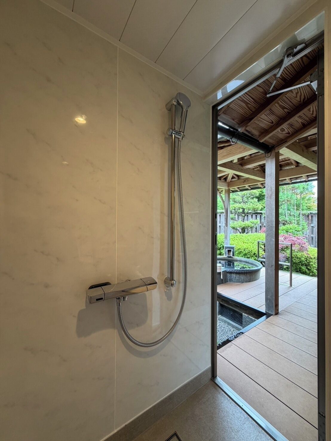Une douche avec un pommeau flexible, un mur carrelé et une terrasse en bois à l’extérieur.