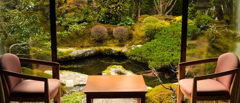 A room with two chairs and a table, a view of a garden with a pond, rocks, and trees.