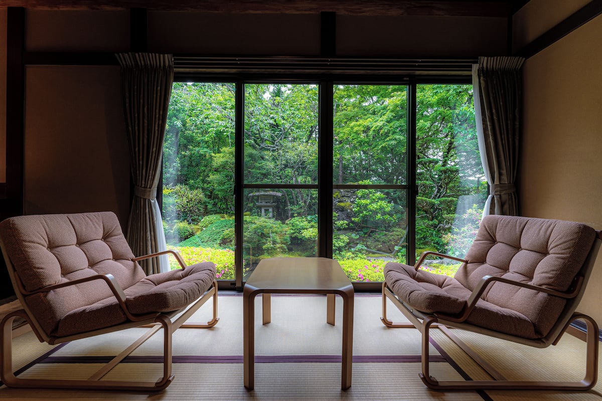 A room with two brown chairs and a small table, large windows with curtains, and a view of greenery outside.