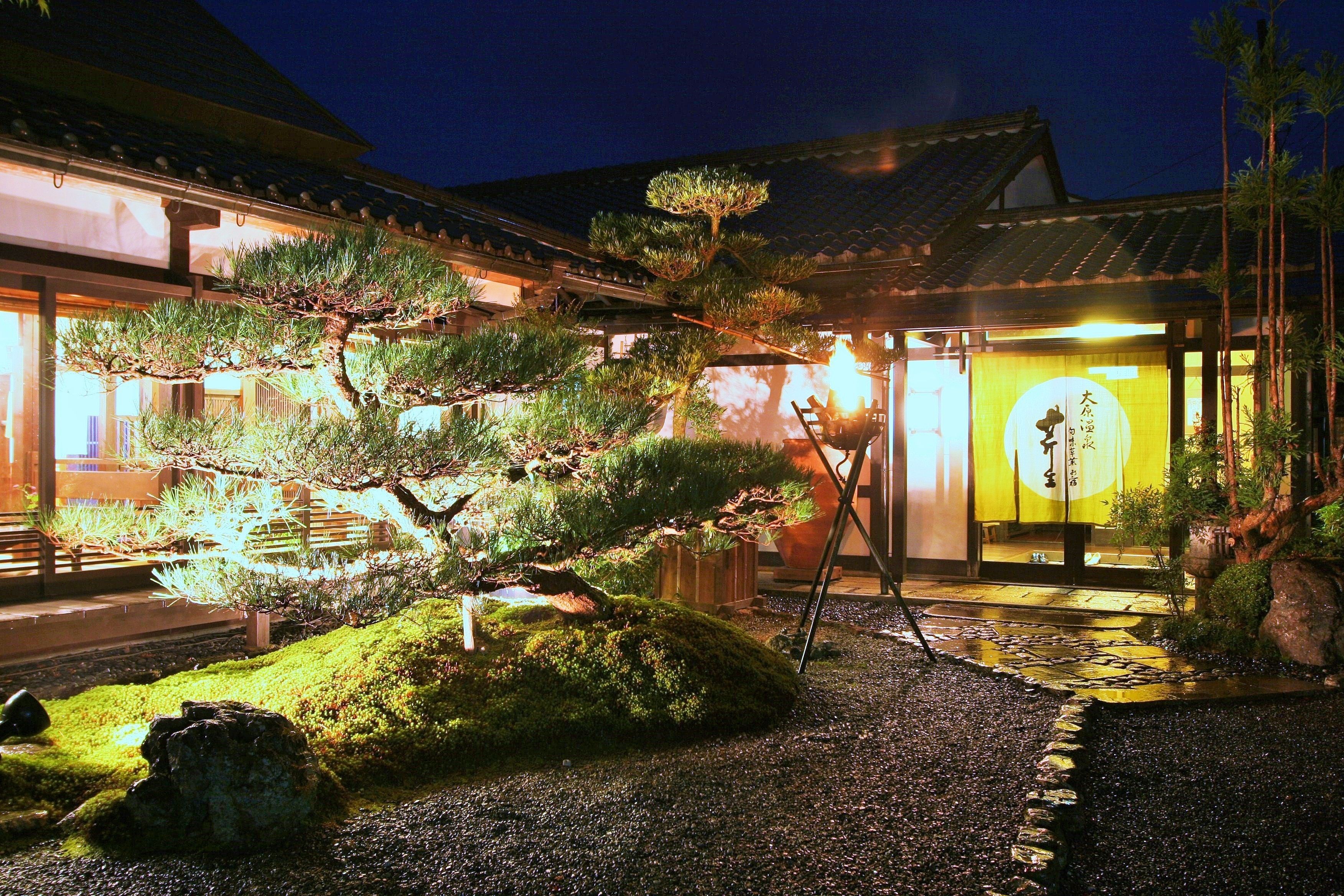 En traditionell japansk byggnad med en välskött trädgård som har tallar, en mossbeklädd sten och en träpromenad.