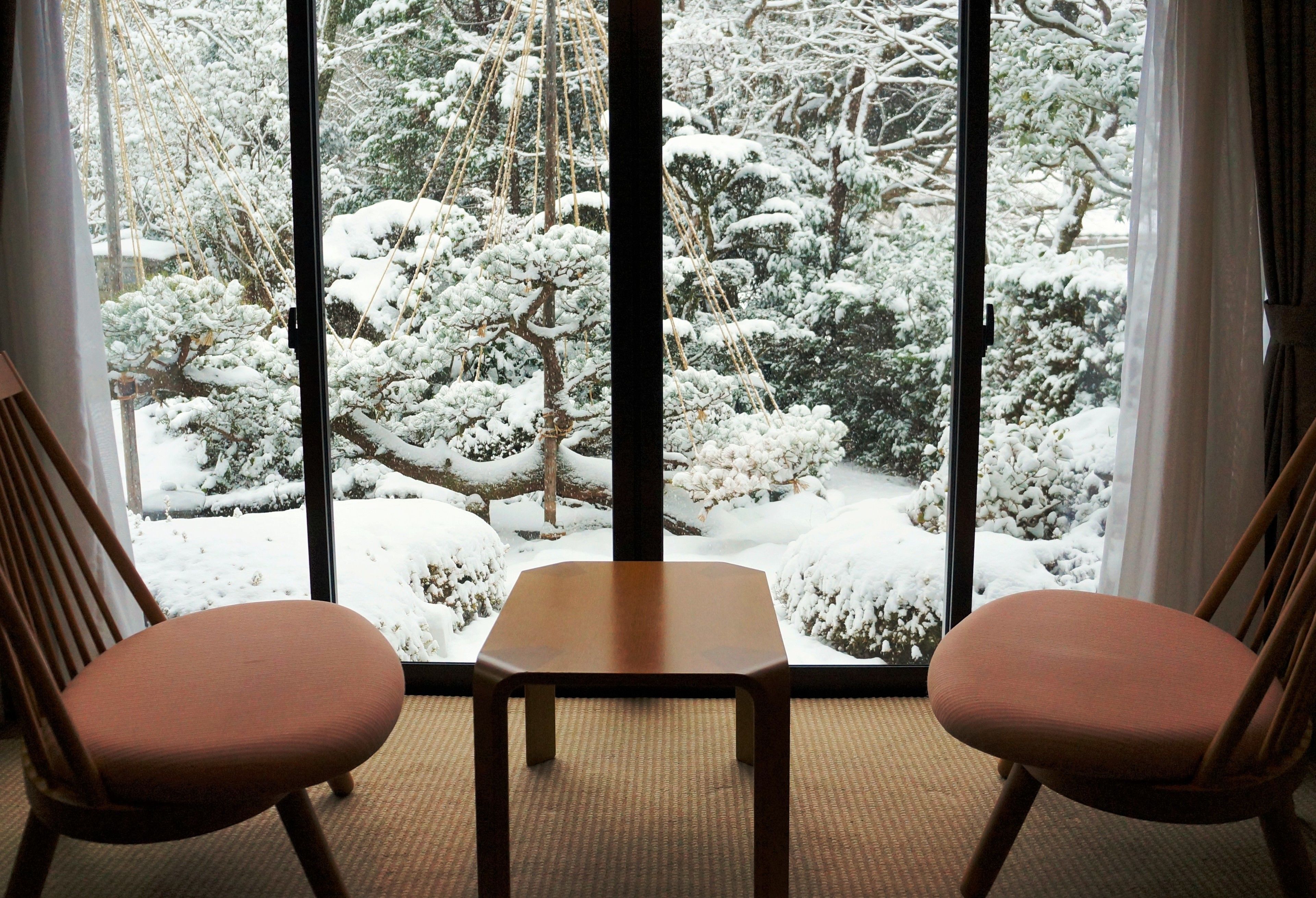 A room with a large window offering a view of a snow-covered garden. There is a wooden table and two chairs in the room.