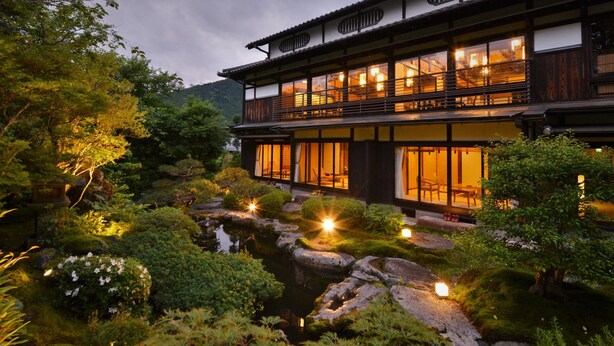 Un edificio tradicional al estilo japonés, con una estructura de madera imponente, varias ventanas y un jardín con rocas y follaje.
