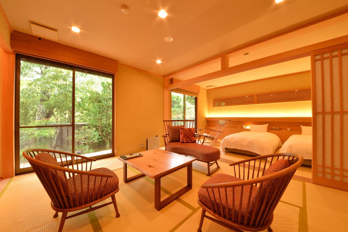 A hotel room with tatami flooring, a traditional Japanese-style sofa, a wooden table, and a bed with a headboard.