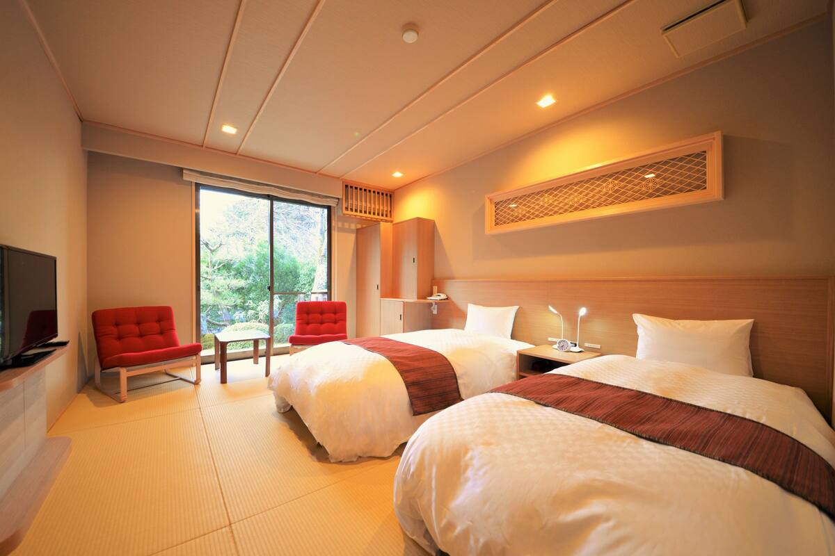 A hotel room with two beds, a TV, a red chair, and a window with a view of greenery.