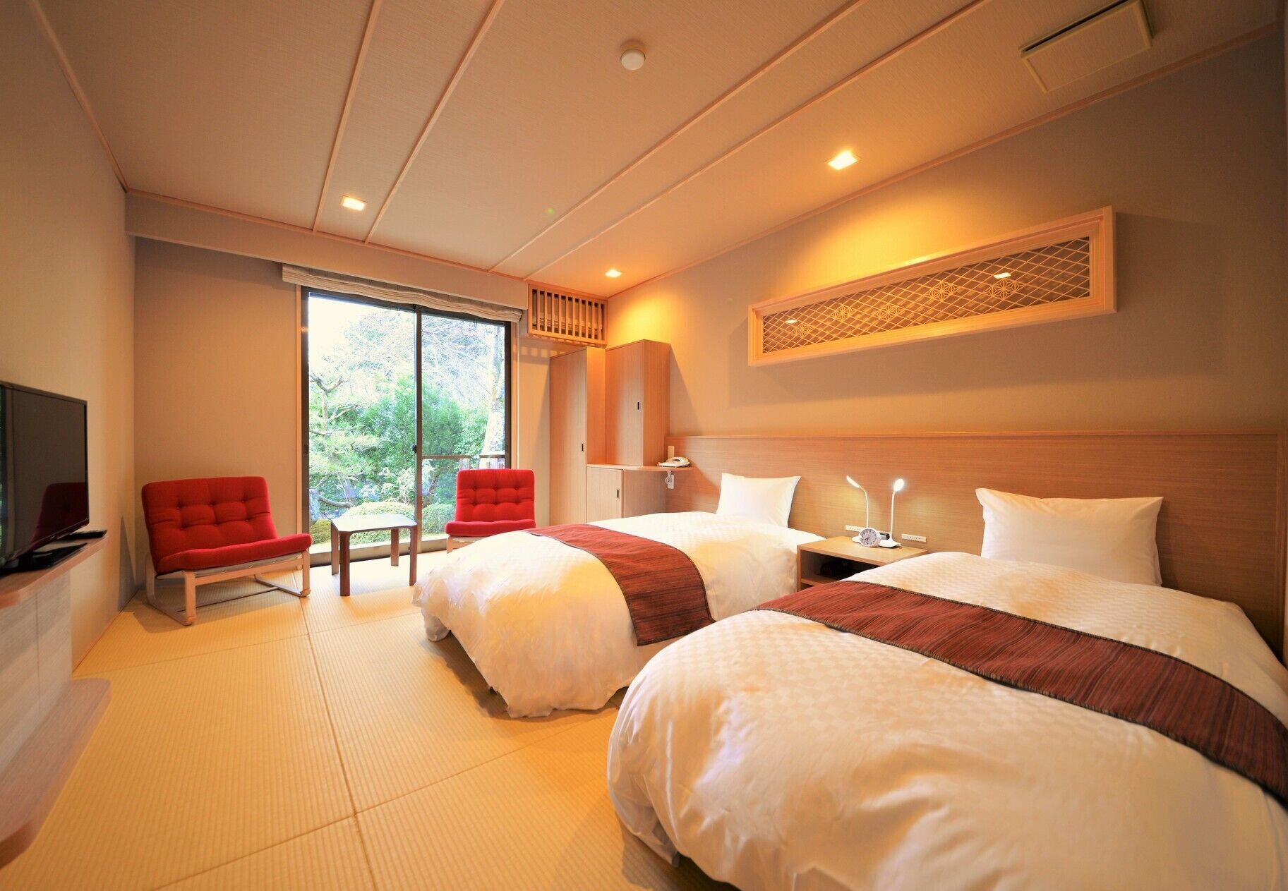 A hotel room with two beds, a TV, a red chair, and a view of greenery.