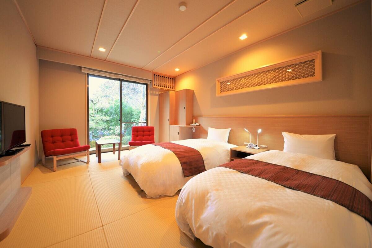 A hotel room with two beds, a TV, a red chair, and a view of greenery.