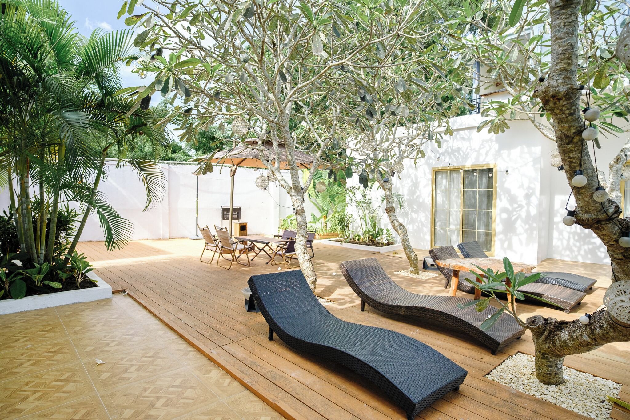 A wooden deck with lounge chairs, a table, and chairs, surrounded by greenery and a white building with large windows.
