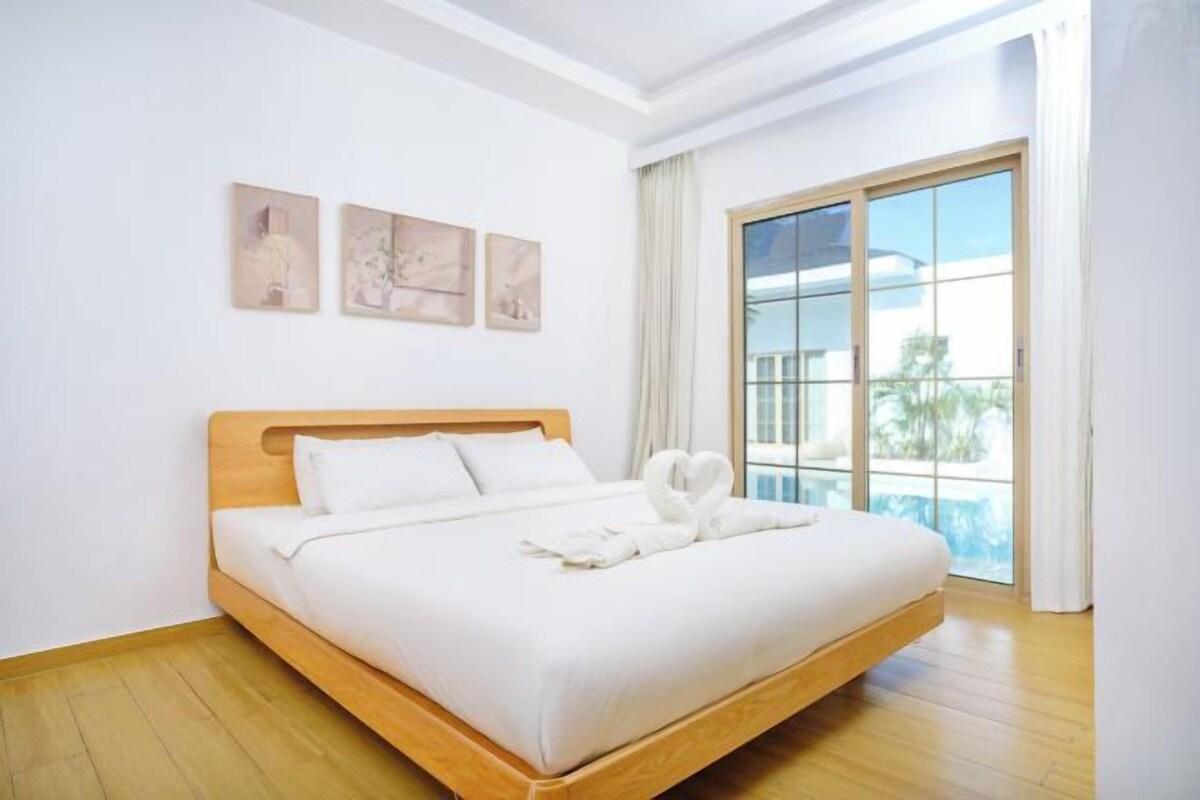A neatly made bed with white linens and a heart-shaped towel arrangement, a wooden headboard, and a large window with a view of palm trees.