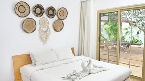 Uma cama bem arrumada com cabeceira de madeira, parede com tapeçarias de tecido e uma grande porta de vidro deslizante que dá para área externa com plantas.