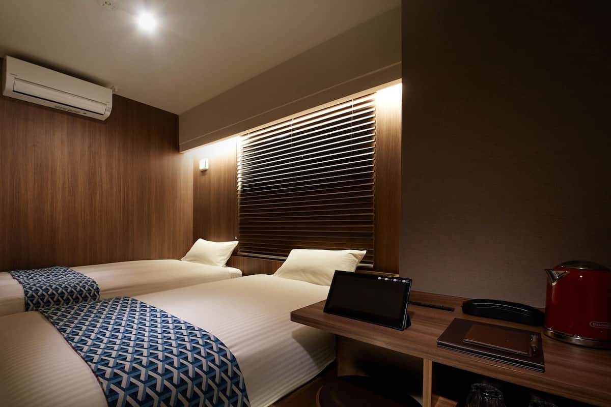 A hotel room with two beds, a desk with a laptop, a red kettle, and a wall-mounted air conditioner.