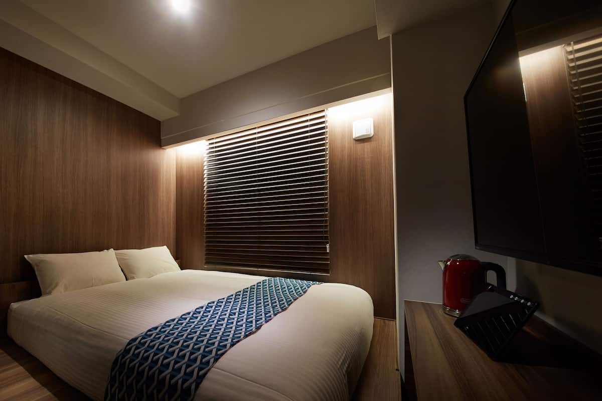 A modern hotel room with a wooden panel wall, a bed with a patterned throw, a bedside table with a kettle, and a large mirror.
